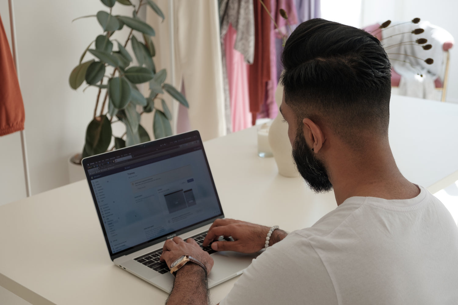 Looking over the shoulder of a man working on his online store on a laptop. There are house plants and clothes hanging in the background. | browse our store to find the perfect items for your enjoyment - storefront for LunaSola Designs